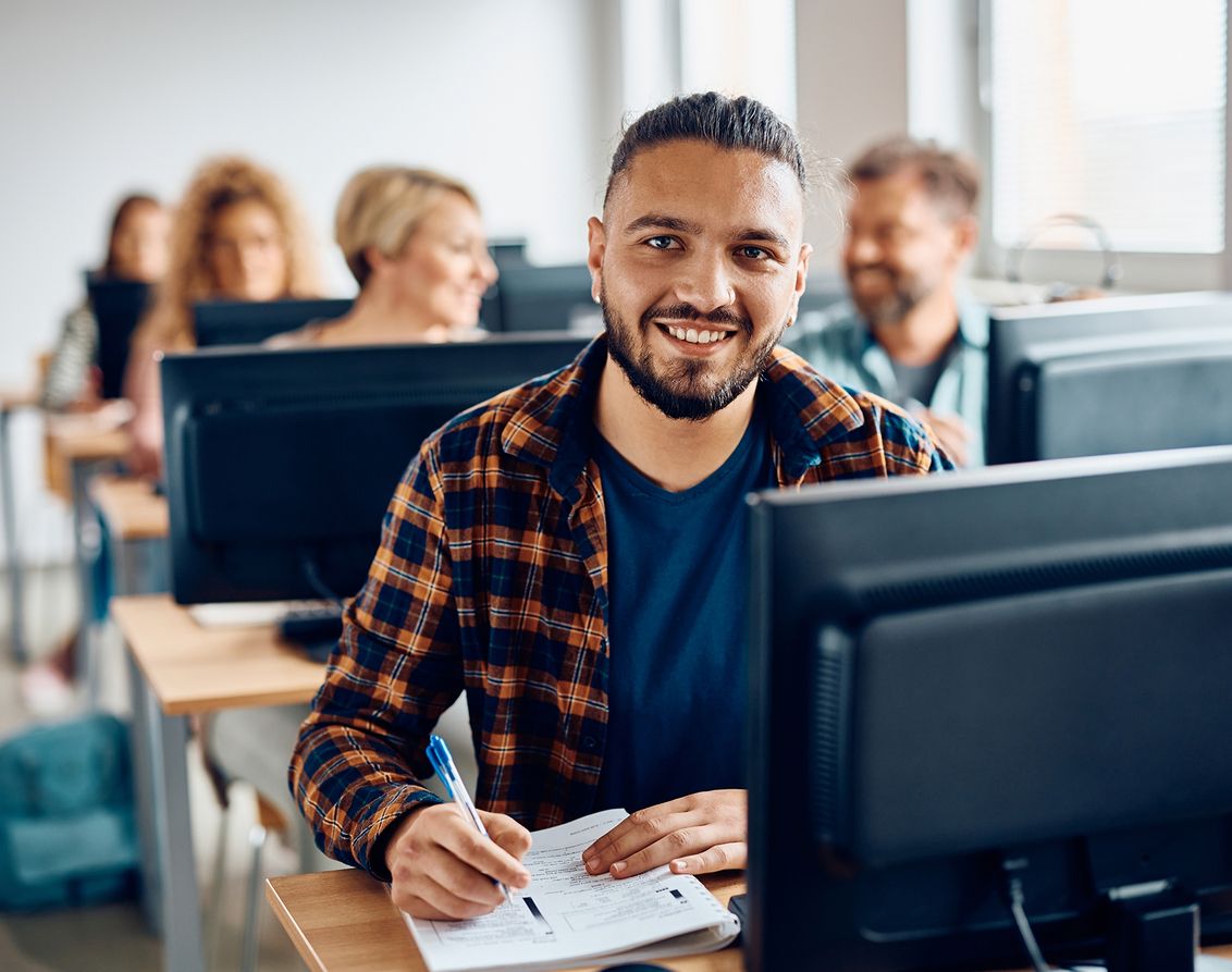 Ein Schüler macht sich an einem Schreibtisch in einem Klassenzimmer Notizen, während im Hintergrund Computer und Mitschüler unscharf zu sehen sind.